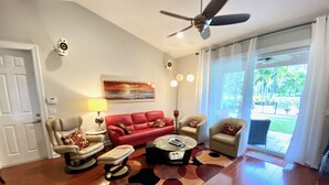 A living room with a red sofa, beige armchairs, a coffee table, and a ceiling fan. - Swans Nest West - Modern & Private, Heated Pool, 3 Brm (Lake worth)