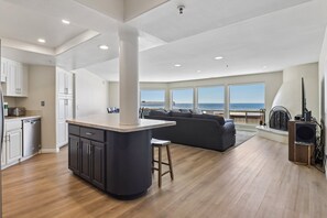 A modern kitchen with a central island, a dining area with a dark sofa, and a large window overlooking the ocean.