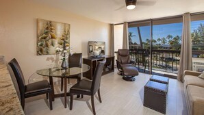 A modern living room with a glass dining table, leather chairs, a sofa, a TV, and a balcony with a view of palm trees.