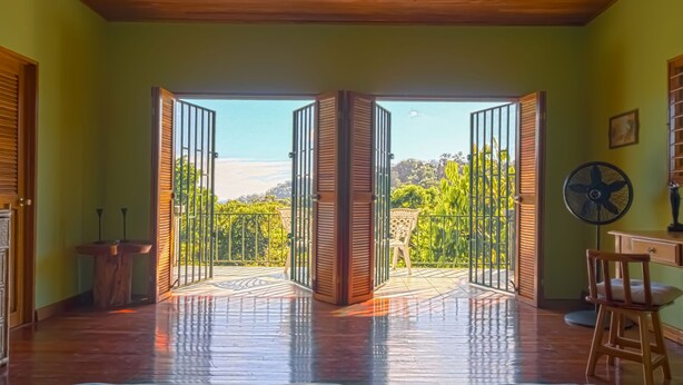 A room with a wooden floor, a desk with a chair, a fan, and a view of greenery through open doors.