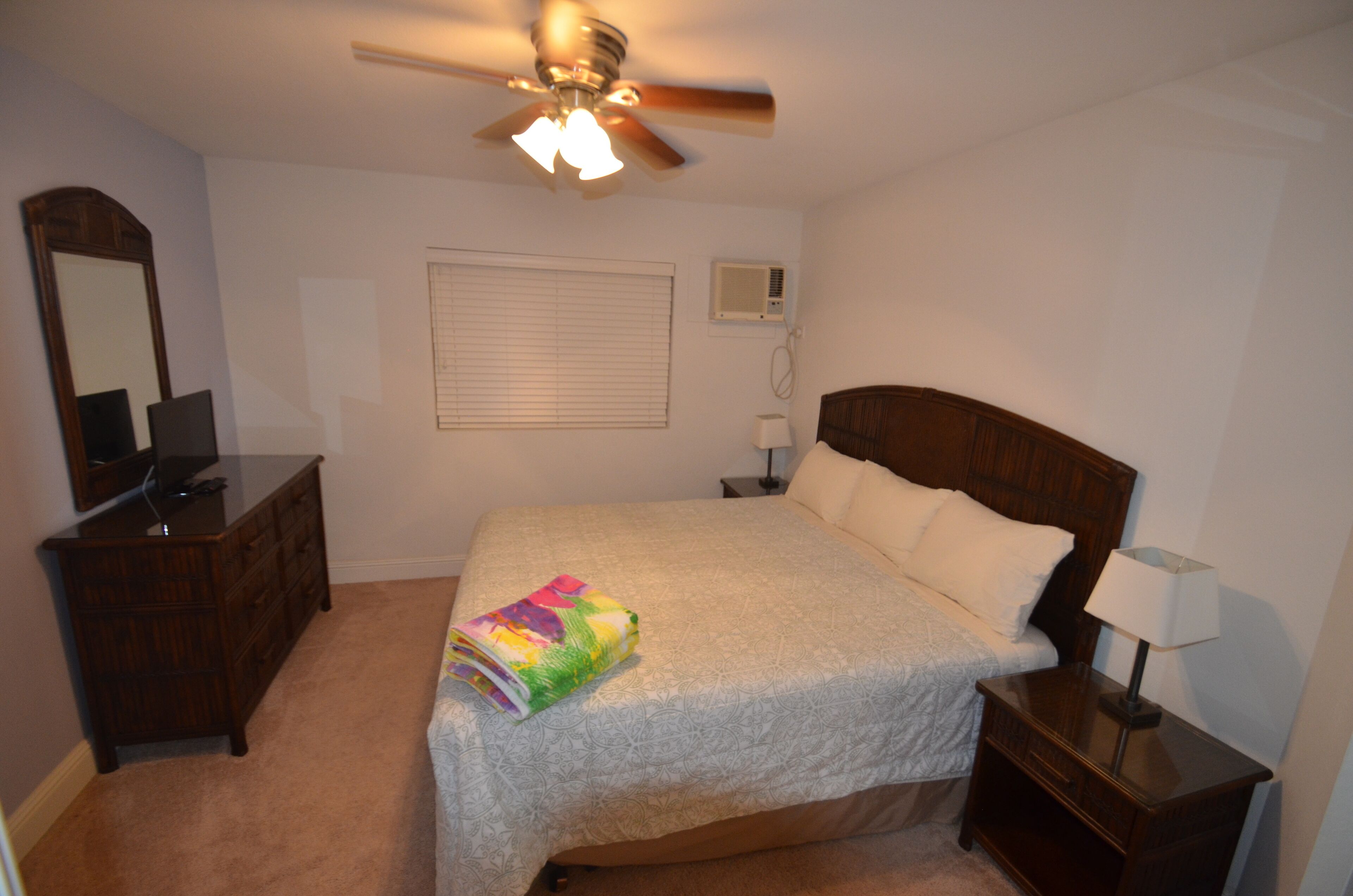 Un dormitorio con cama, cómoda, mesita de noche, ventilador de techo, ventana con persianas y unidad de aire acondicionado.