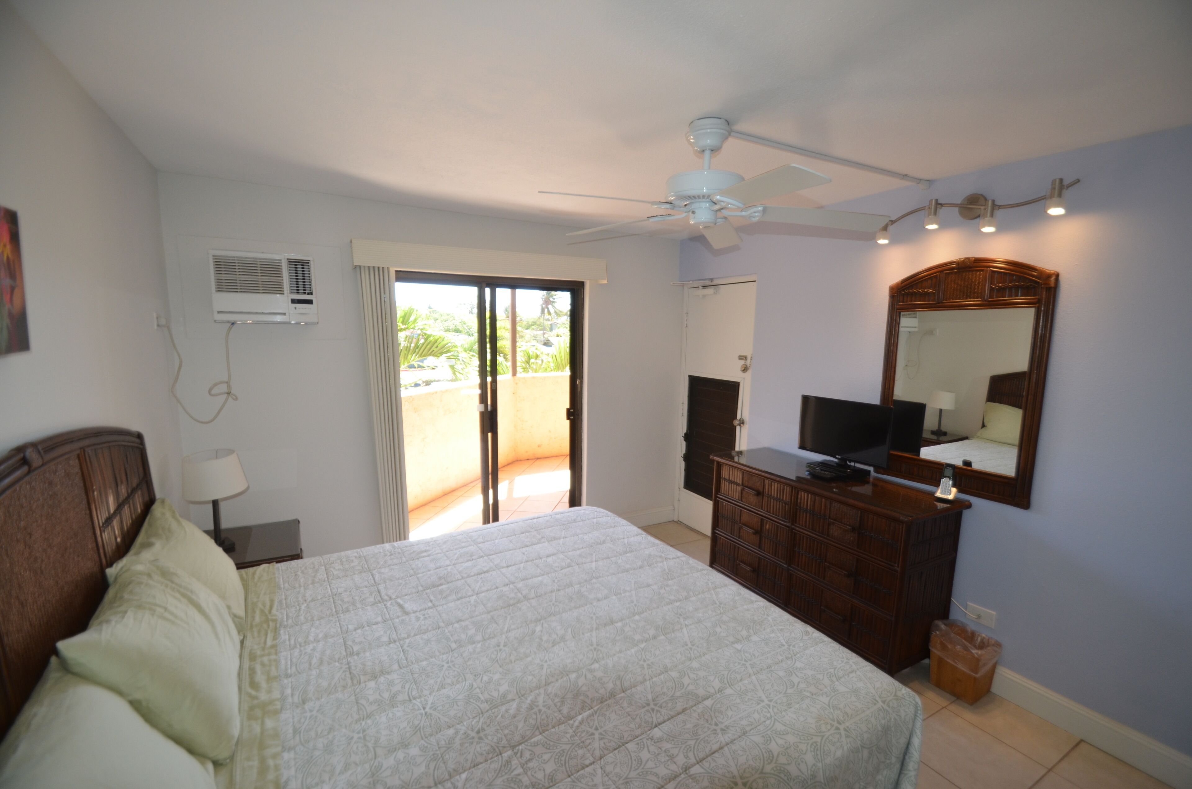 A bedroom with a bed, a dresser, a ceiling fan, a TV, and a window with a view of the outdoors.