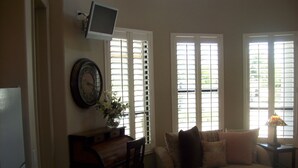 A living room with a sofa, a coffee table, a TV mounted on the wall, a clock, and a window with blinds. - Biltmore Golf and Resort (just steps away from the fabulous Biltmore Hotel) (Phoenix)