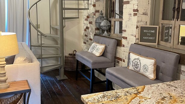 A modern living room with a staircase, a sofa, a coffee table, and a decorative wall with a 'Welcome to Natchtez!' sign. - Charming 2 bedroom condo in the heart of beautiful Natchez, MS. (Natchez)