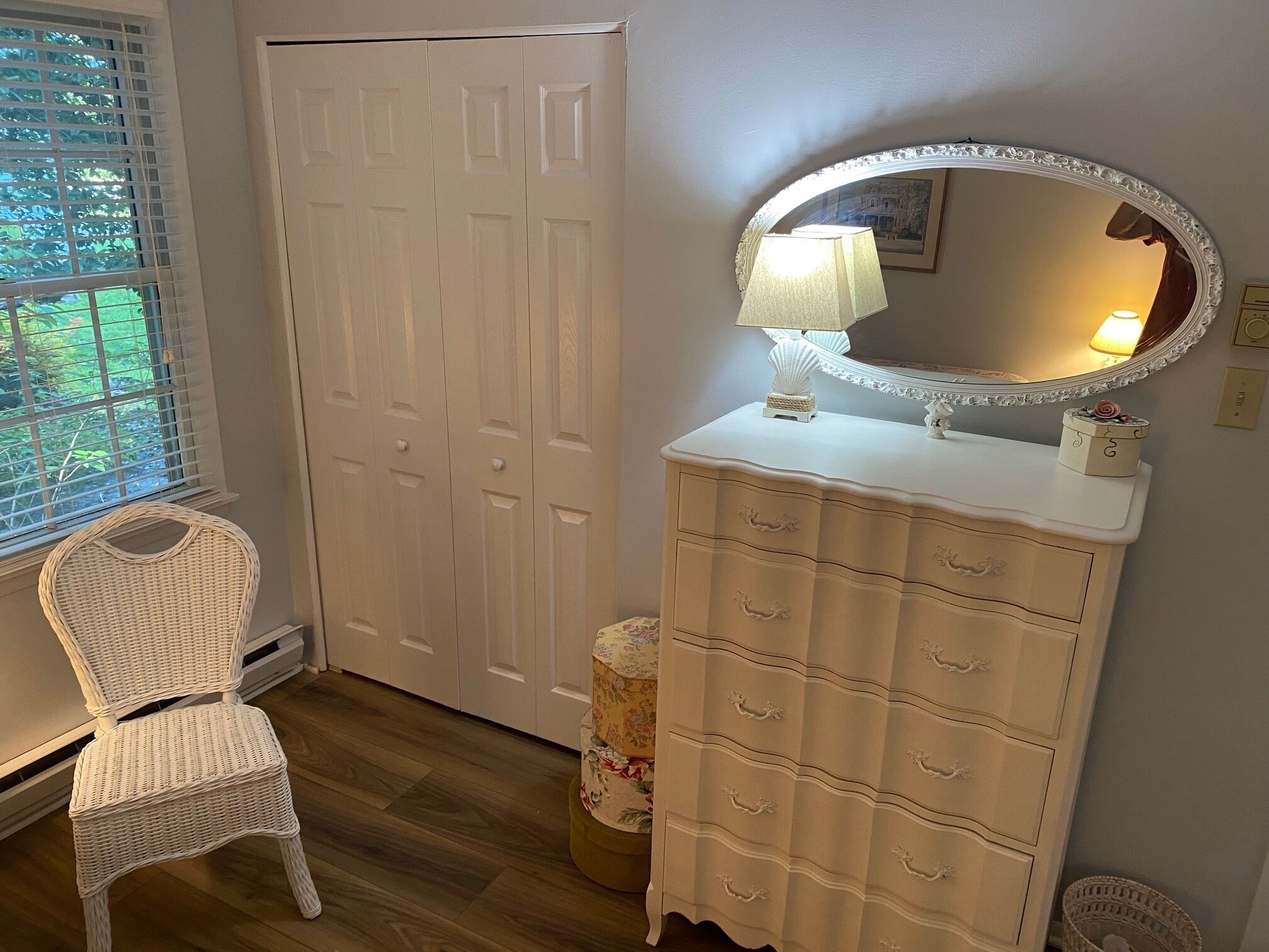 Une pièce avec une commode blanche, un fauteuil rond en osier, une porte blanche fermée et une fenêtre avec des stores.