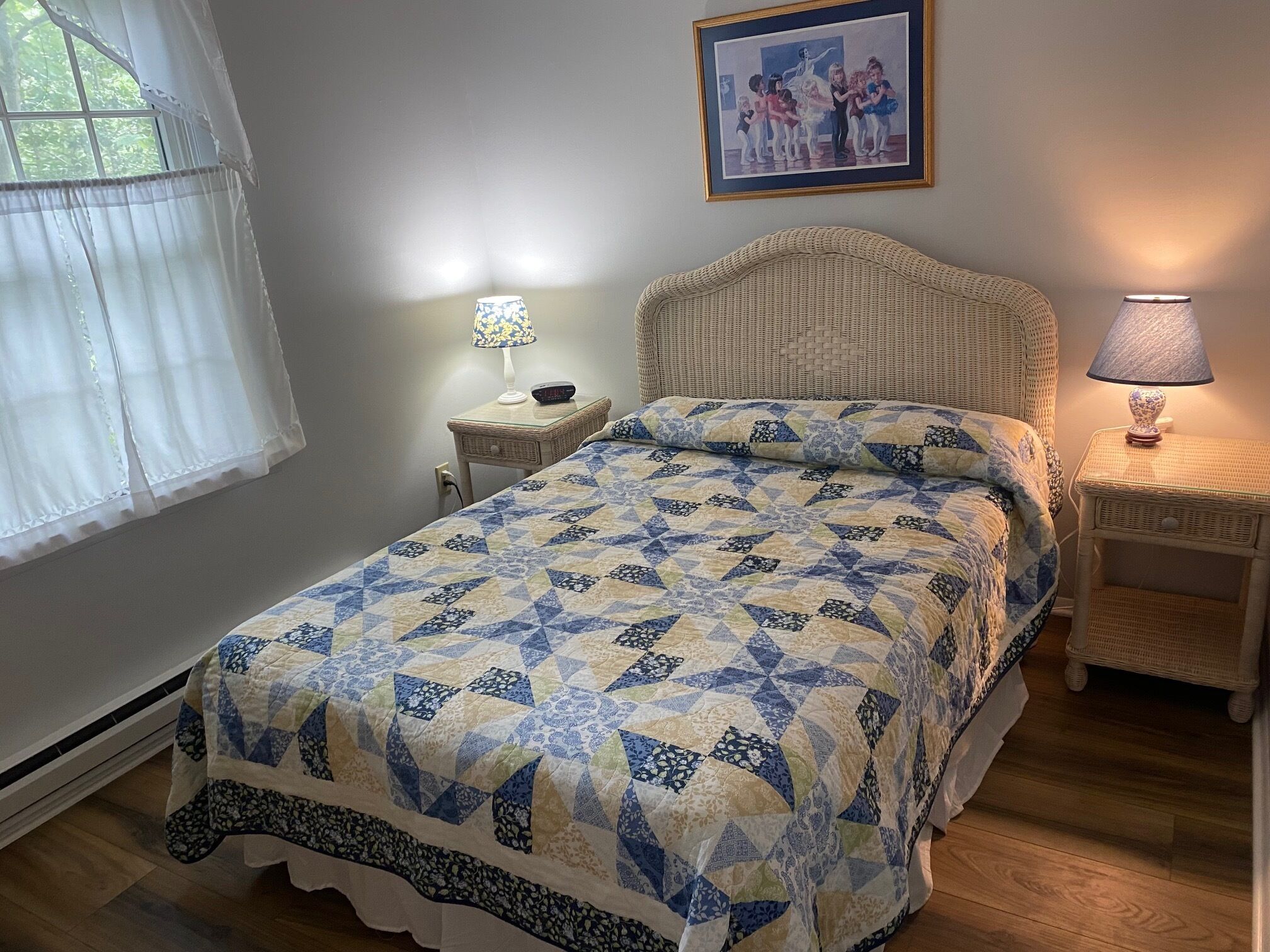 Ein Schlafzimmer mit einem Bett, einem Nachttisch, einer Lampe, einem gerahmten Bild an der Wand und einem Fenster mit Vorhängen.