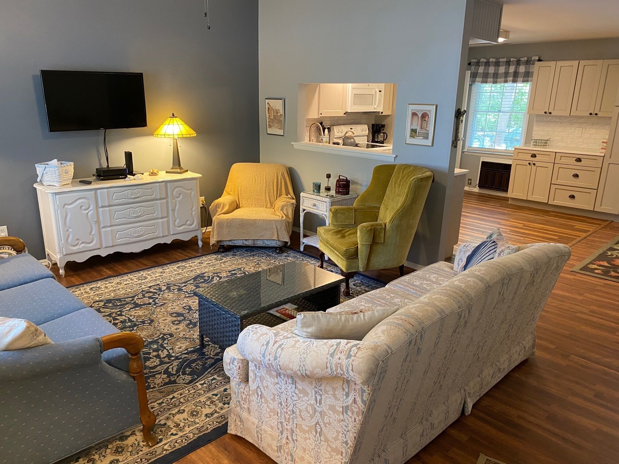 A living room with a sofa, armchair, and coffee table, a television, and a kitchen in the background.