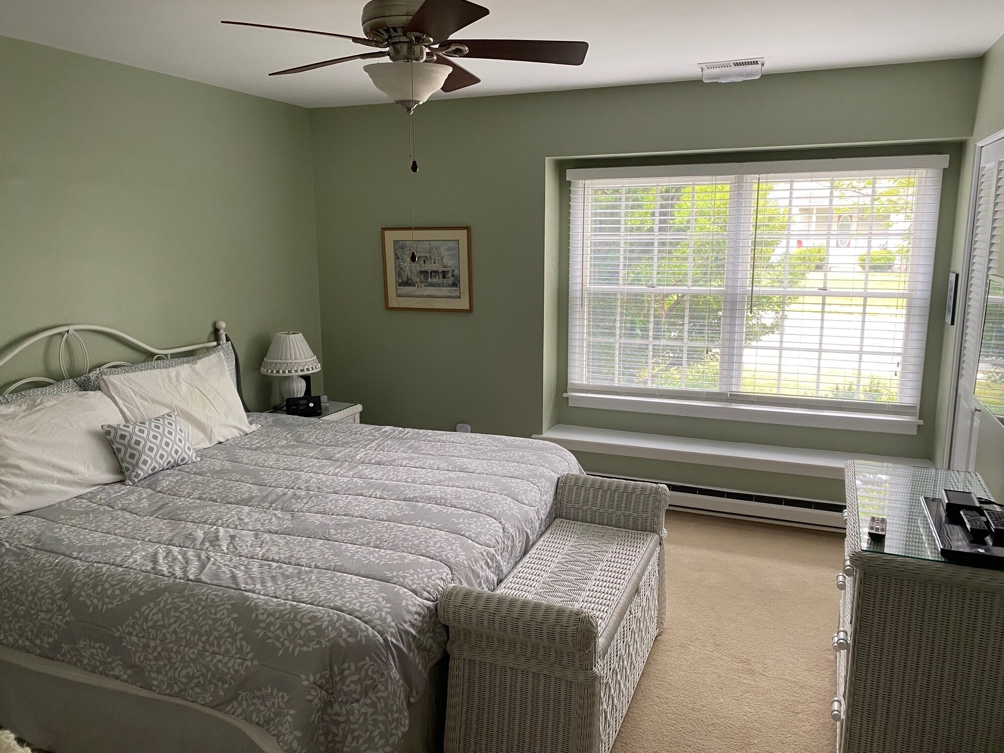 A bedroom with a bed, a ceiling fan, a window with blinds, a bedside table with a lamp, and a framed picture on the wall.