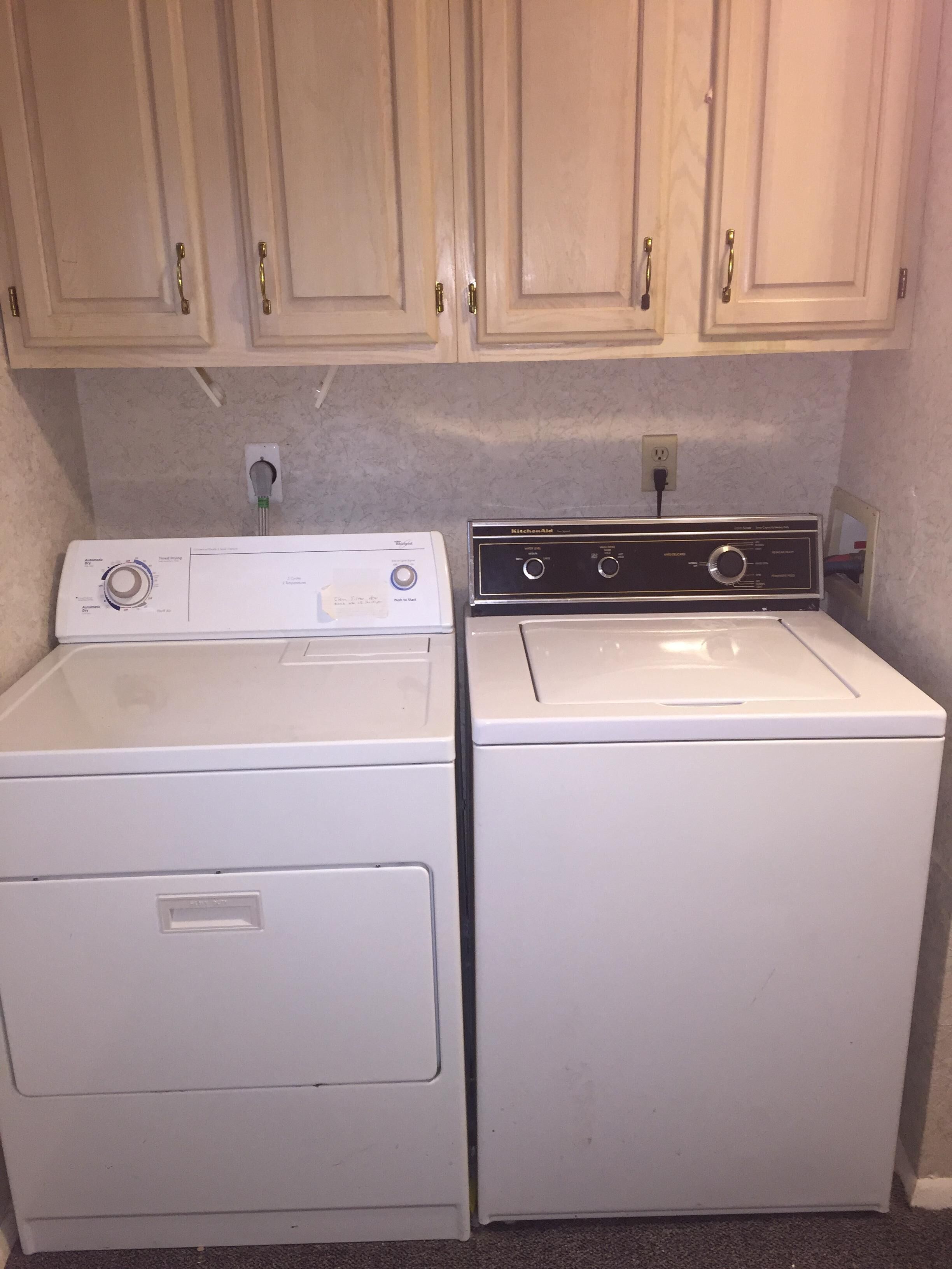 A laundry room with two white washing machines and a dryer, wooden cabinets above, and a countertop with a power outlet.