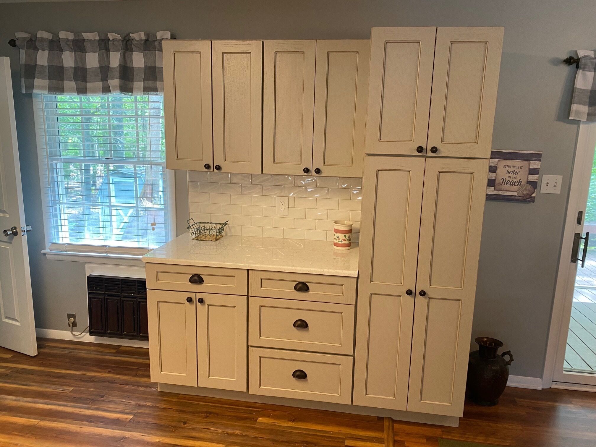 Uma cozinha com armários claros, uma janela com persianas e um letreiro decorativo na parede.