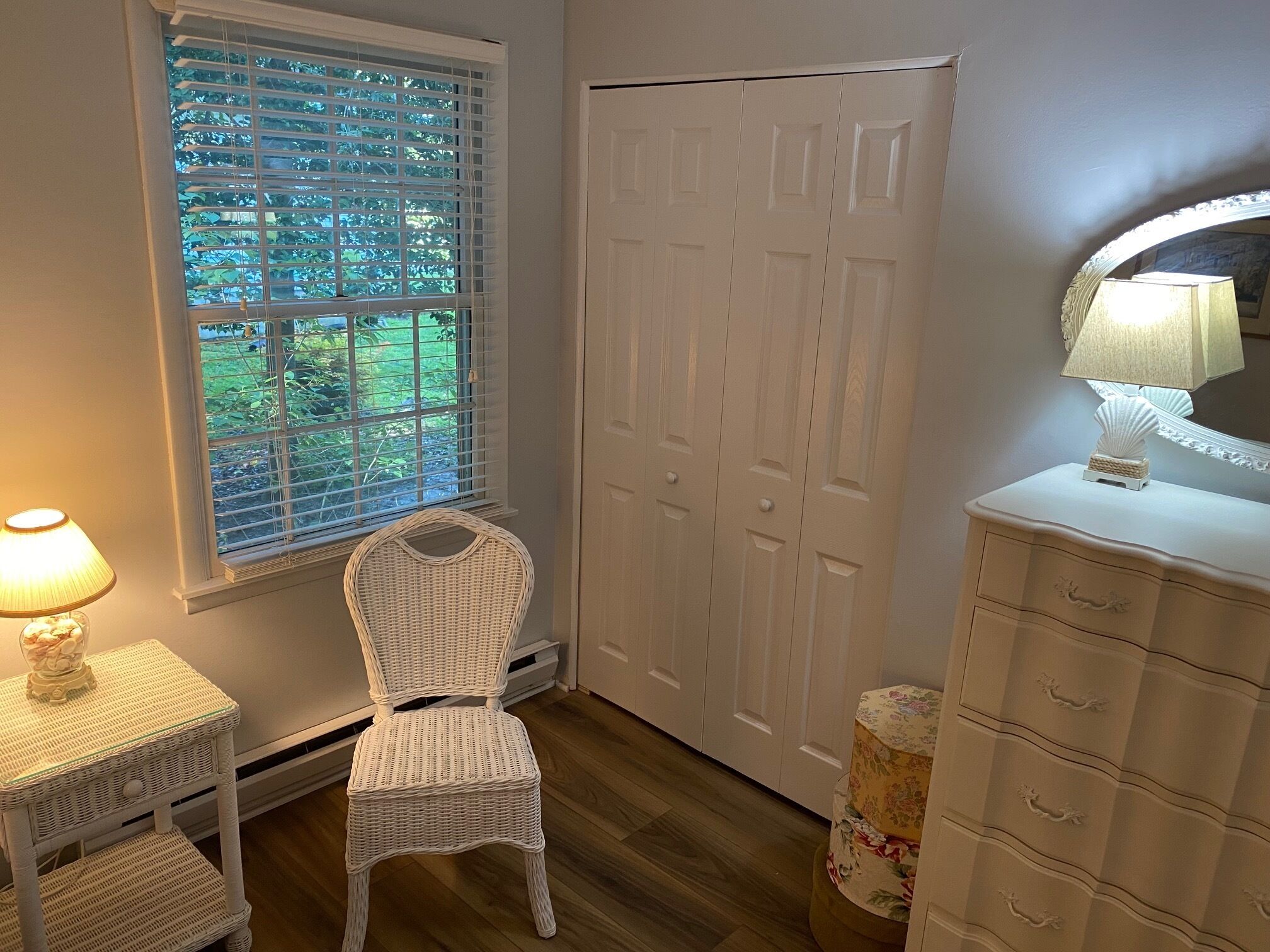 A room with a wicker chair, a white dresser, a mirror, and a window with blinds.