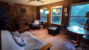 A cozy room with a wooden floor, a couch, a chair, a small table, and a window with a view of trees.