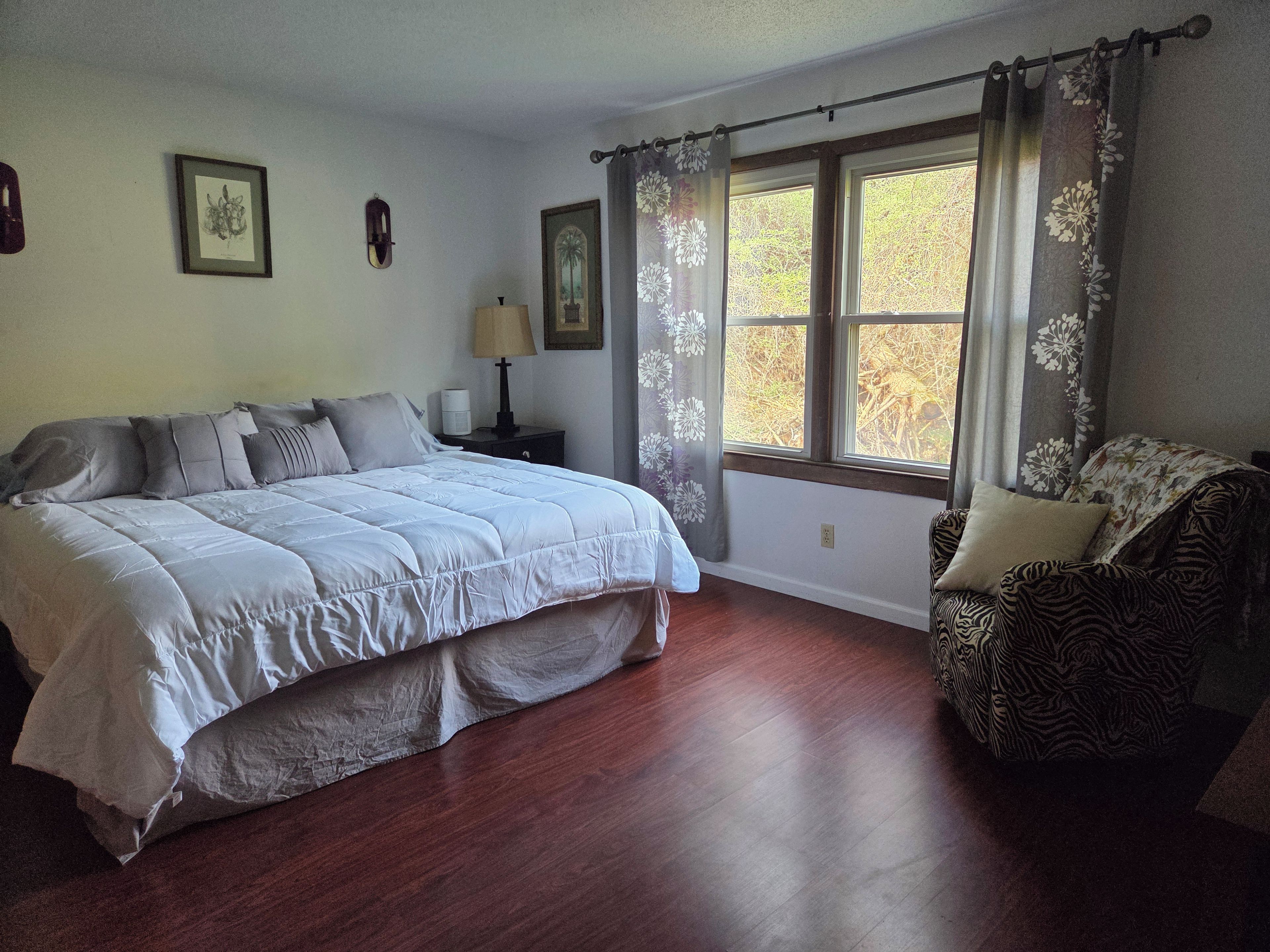 A bedroom with a large bed, a nightstand with a lamp, a window with curtains, and a patterned armchair.