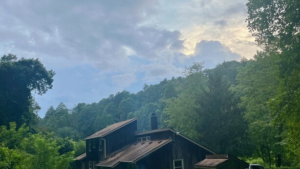 A rustic wooden house with a chimney, surrounded by lush greenery and a grassy yard.