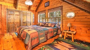Un dormitorio de cabaña de madera acogedor, con una cama grande, una mesita de noche con lámpara y una ventana con vistas a árboles.