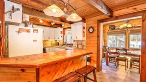 Interior de una cabaña de madera con una zona de cocina que incluye una barra de madera, taburetes y una mesa de comedor con sillas.