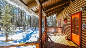 Una cabaña de madera con un porche, una puerta roja y vistas a un bosque nevado.