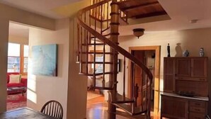 Un escalier en colimaçon en bois menant à l’étage supérieur, un coin repas avec une table et des chaises en bois, et un espace cuisine avec des placards en bois.