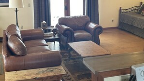 Una sala de estar con un sofá de cuero marrón, un sillón a juego, una pequeña mesa de madera y una cama con una manta gris.