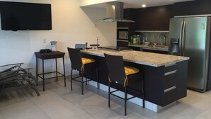 Una cocina moderna con una isla central, taburetes de bar, un televisor de pantalla plana y un refrigerador de acero inoxidable.