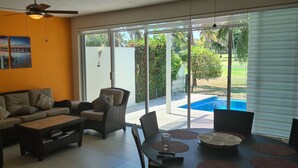 Una sala de estar con sofá, sillas, una mesa de centro y una mesa de comedor con individuales. Hay un ventanal con vistas a una piscina y vegetación exterior.