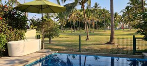 Zona de piscina con una sombrilla verde, una maceta blanca y vistas a un campo de golf con palmeras.