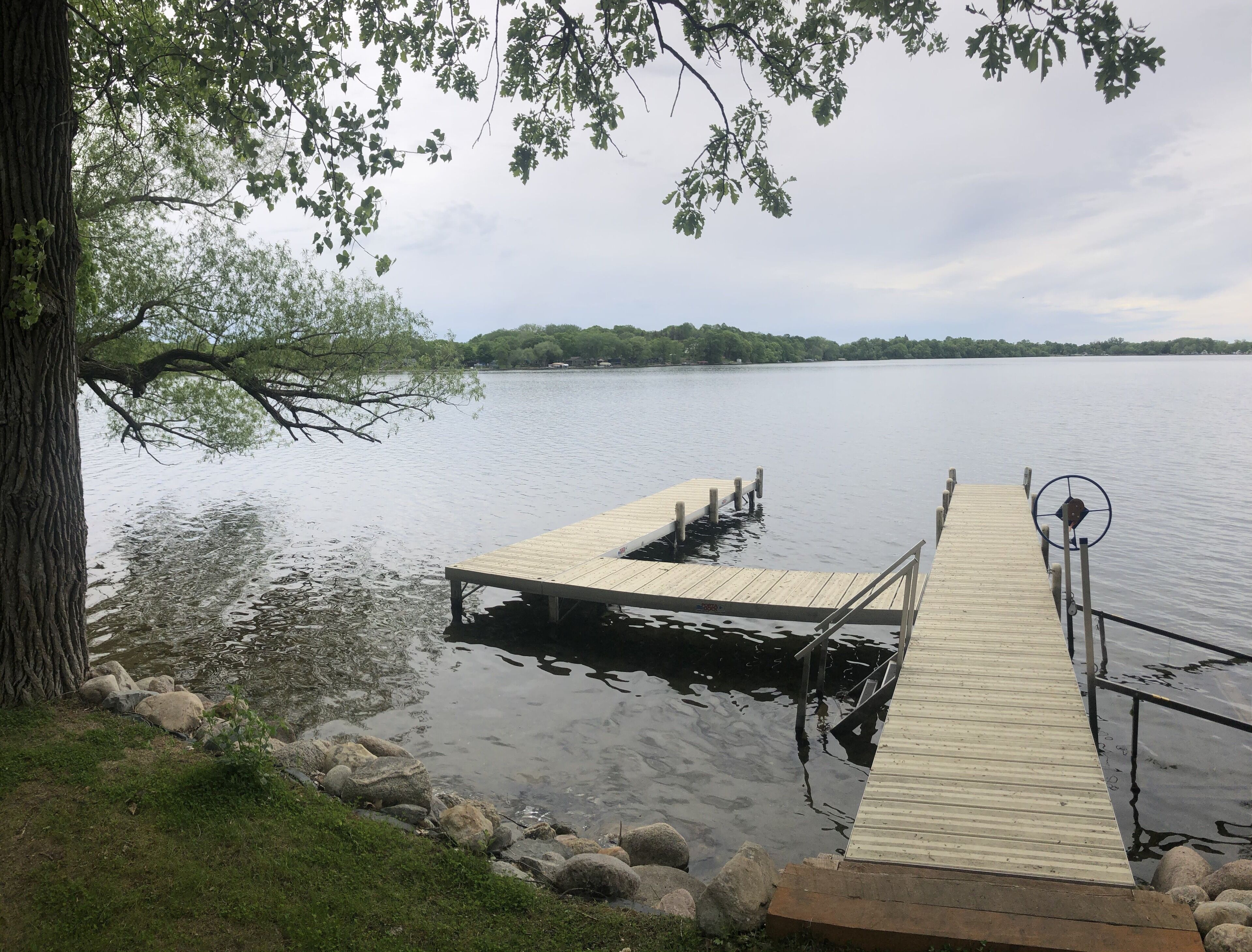 A wooden dock extending into calm water, surrounded by rocks and grass, with a tree on the left and a clear view of the water and distant shoreline.