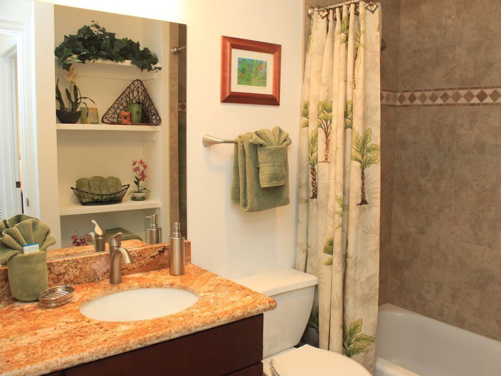 Une salle de bain avec un lavabo sur un plan de travail en granit, des toilettes, un rideau de douche orné d’un motif de feuilles et un tableau encadré au mur.