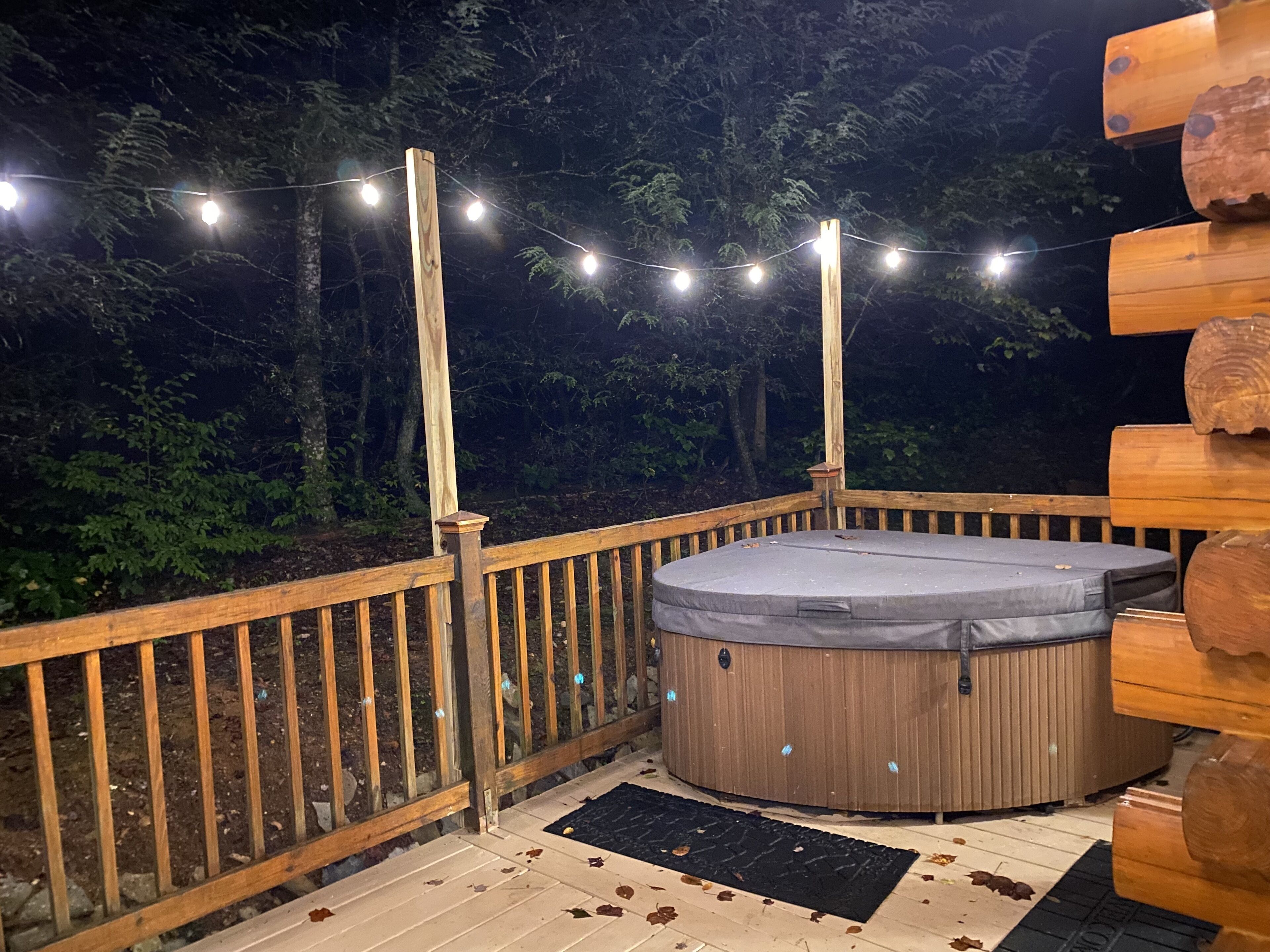 Una terraza de madera con jacuzzi, luces colgantes y un cerco de madera.