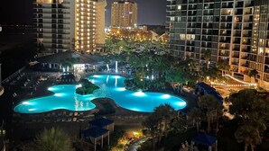 A nighttime view of a hotel with a large, illuminated swimming pool surrounded by palm trees and lounge chairs.