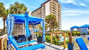 A wooden deck with blue lounge chairs and a canopy, overlooking a pool and palm trees, with a tall building in the background.