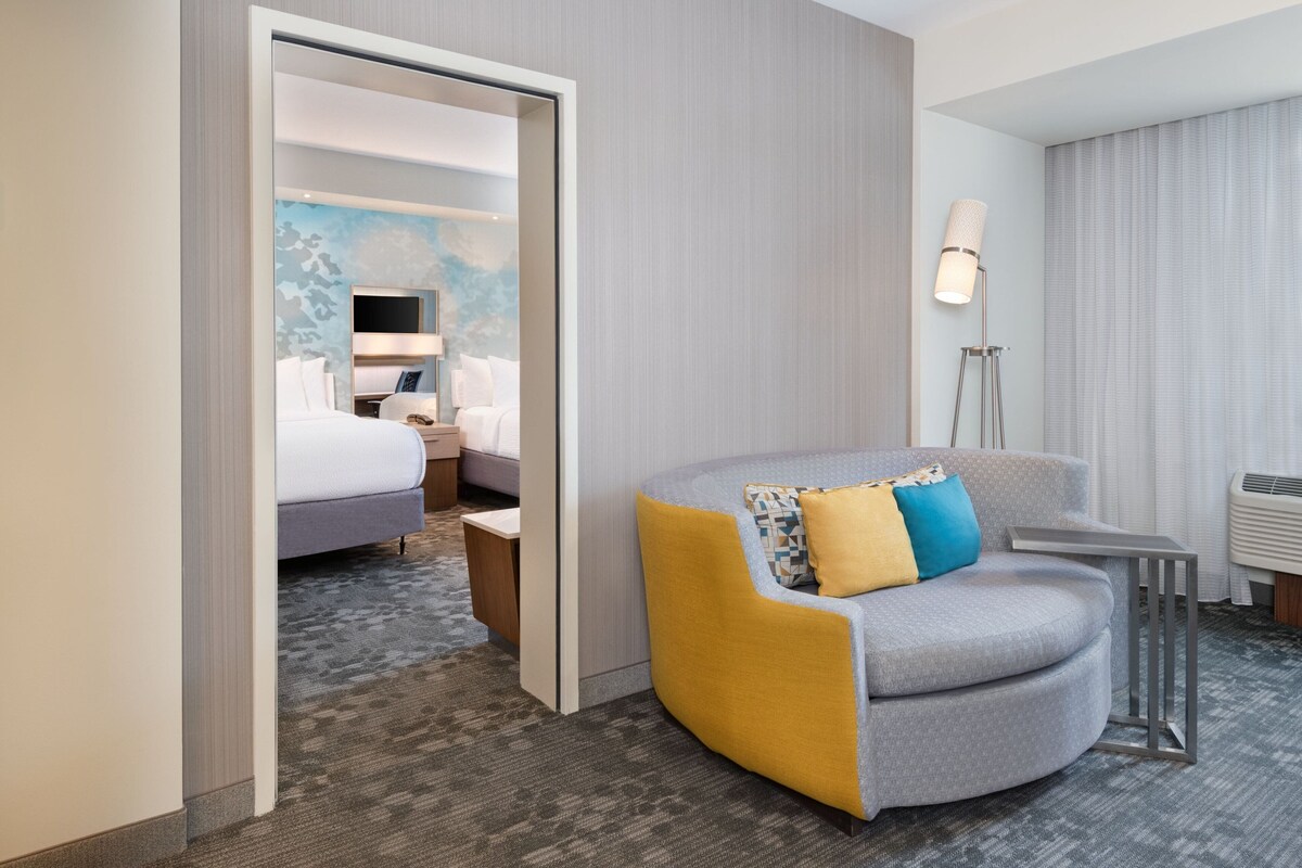 A hotel room with a bed, a seating area with a chair and a small table, a floor lamp, and a wall with a patterned design.
