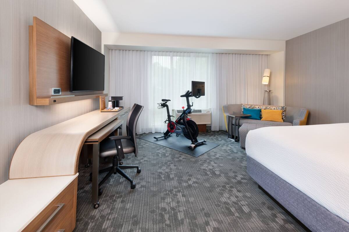Habitación de hotel con escritorio, silla, cama, bicicleta estática y televisor montado en la pared.