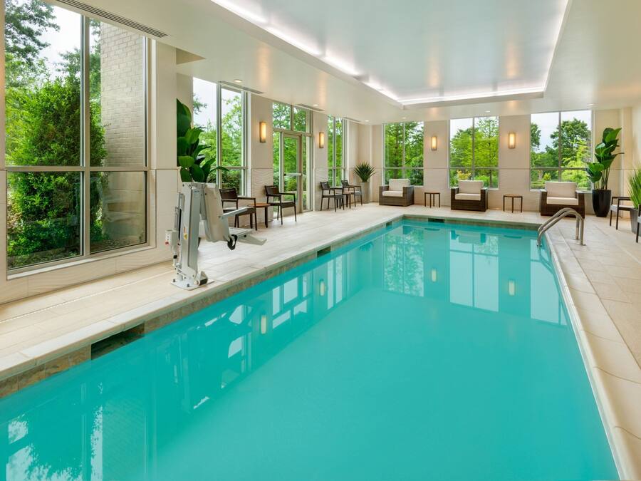 Una piscina interior con paredes de vidrio transparente, rodeada de sillones y mesas, y grandes ventanales con vistas a zonas verdes.