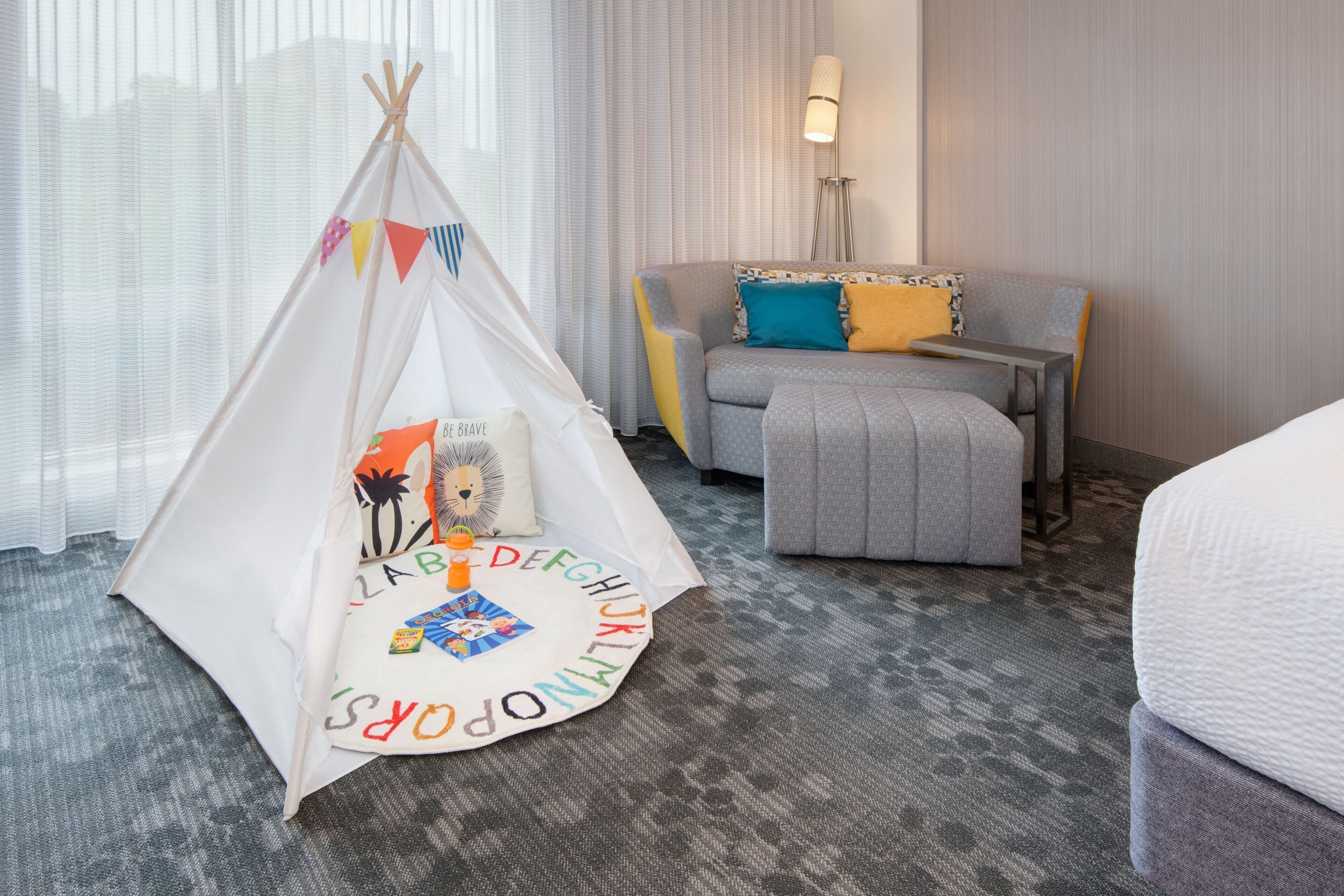 A hotel room with a bed, a grey sofa, a small table, a lamp, and a white tent with colorful letters.