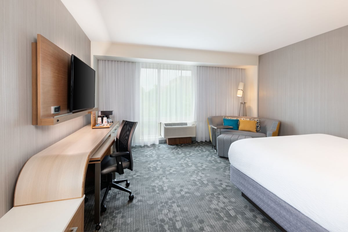 Habitación de hotel con cama, escritorio con silla, televisión montada en la pared y ventana con cortinas.