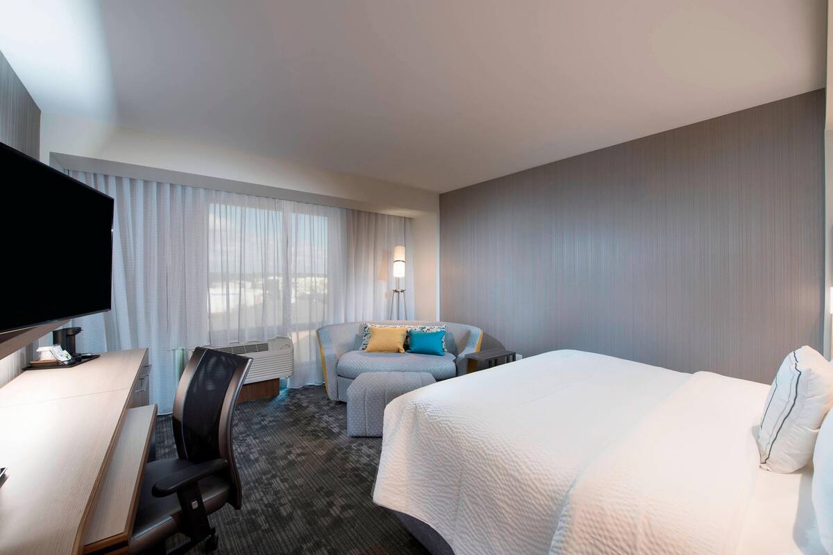 Habitación de hotel con cama, escritorio, silla, televisor y ventana con cortinas.