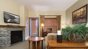 A cozy room with a stone fireplace, a wooden table, a comfortable chair, and a large window with blinds. - Soaring Eagle #406:  2 Bedrooms, 2 Full Baths:  Slope Side,  Hot Tubs. Underground Parking. (Snowshoe)