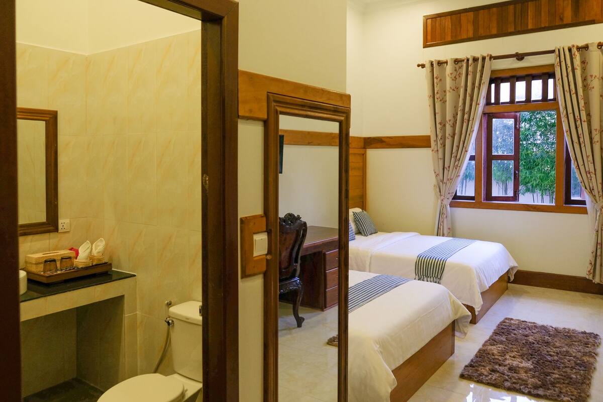A hotel room with two beds, a wooden desk, a mirror, and a window with curtains.