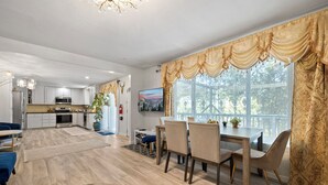Ampla sala de estar com mesa e cadeiras, cozinha ao fundo e uma grande janela com cortinas.