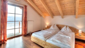 Ein Schlafzimmer mit Holzbalkendecke, einem großen Fenster mit Vorhängen, einem Bett mit weißer Bettwäsche und einem Nachttisch mit Lampe.