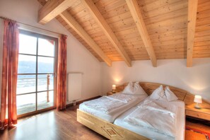 Une chambre avec un plafond en bois, une grande fenêtre avec des rideaux, un lit recouvert de draps blancs et une table de chevet avec une lampe.