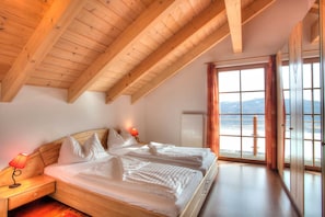 A bedroom with a wooden ceiling, a bed with white bedding, a nightstand with a lamp, and a window with a view of the mountains.