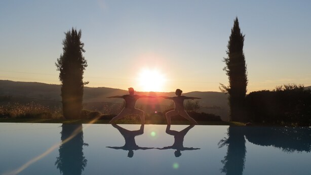 Dos personas realizando una postura de yoga sobre un estanque reflectante, con un atardecer de fondo.