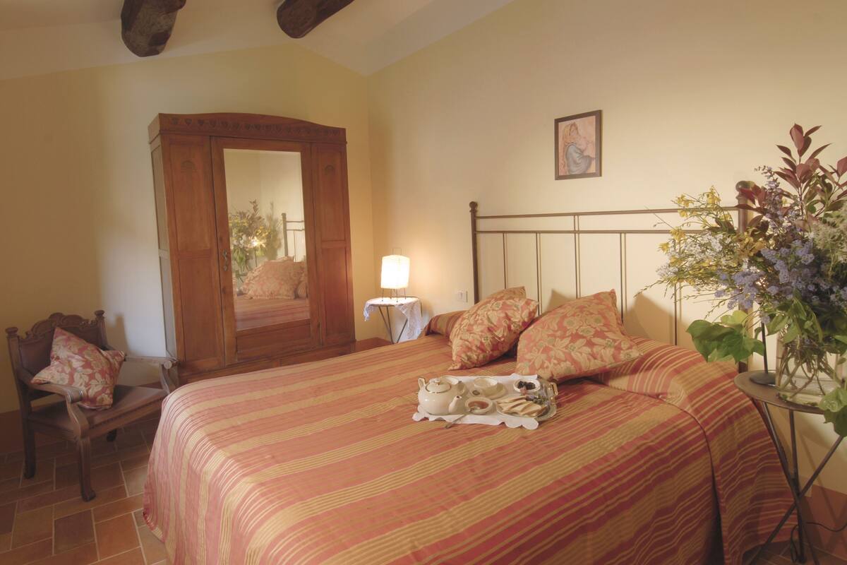 A bedroom with a large bed, a wooden wardrobe, a small bedside table with a lamp, and a potted plant.