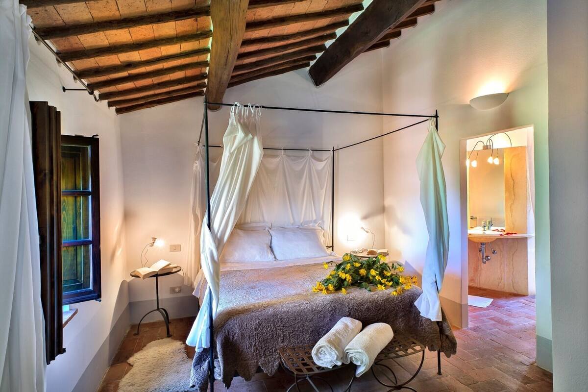 A bedroom with a four-poster bed, a bedside table, a lamp, and a bathroom with a sink and mirror.