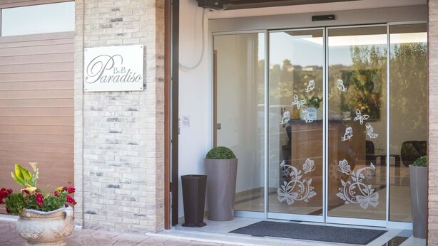 Entrada a un restaurante con un letrero que dice "B&B Paradiso", una puerta de cristal decorada con pegatinas de mariposas y plantas en macetas afuera.