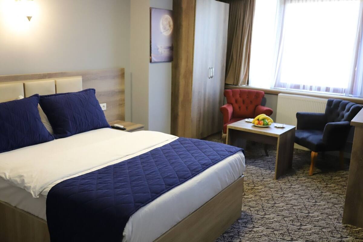 A hotel room with a large bed, a desk with a chair, a red armchair, and a window with curtains.