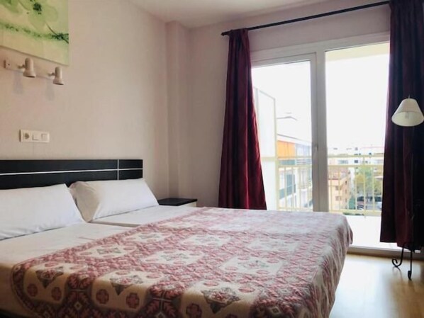 Una habitación de hotel con cama, mesita de noche, ventana con cortinas y vistas a edificios.