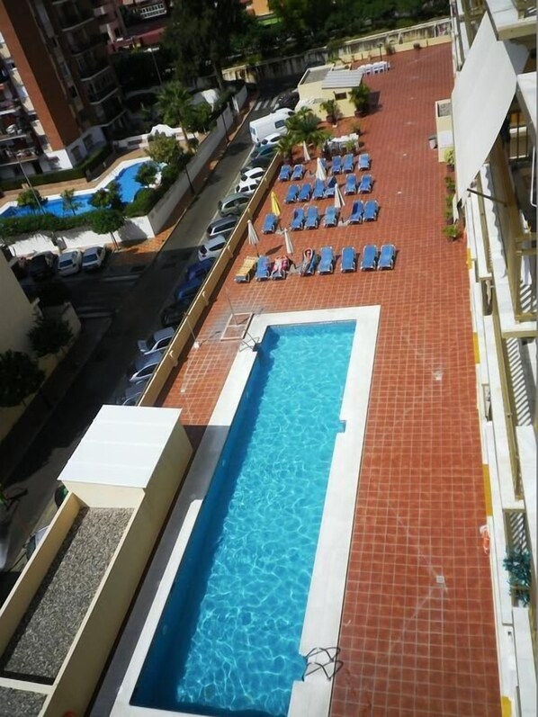 A rooftop pool with blue water, surrounded by lounge chairs and umbrellas, with buildings and a street in the background.
